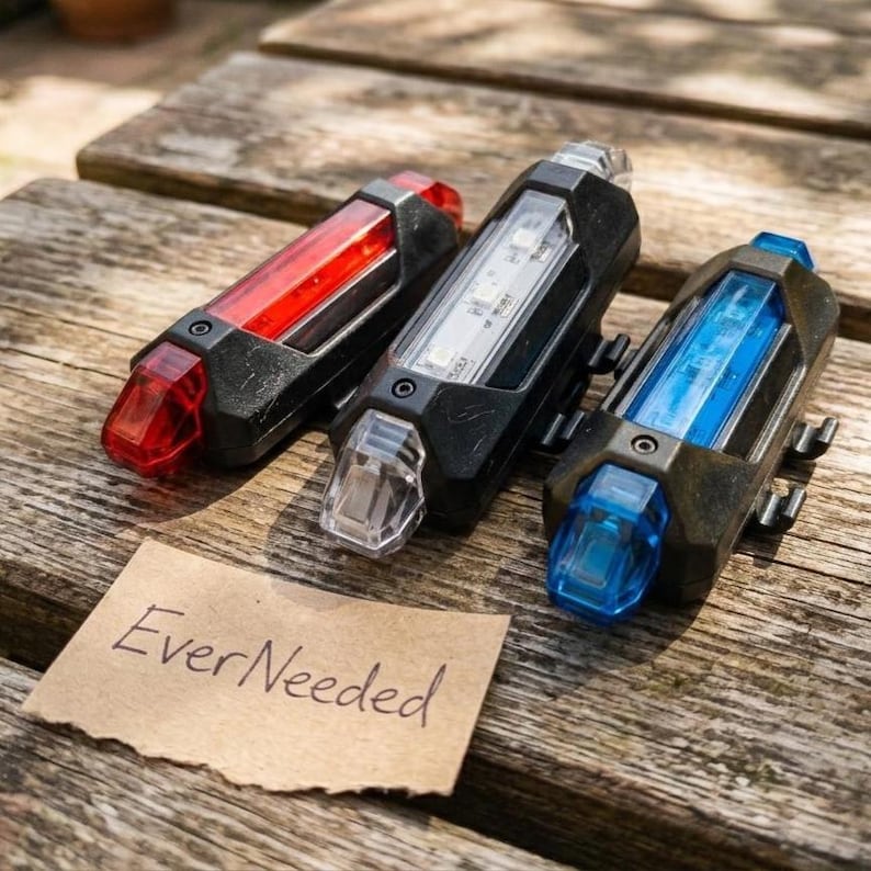 May include: Three bicycle lights with black casings and clear, red, white, and blue lenses are displayed on a wooden surface. The lights have a rectangular shape and are designed for visibility. A small piece of paper with the text "EverNeeded" is also present.