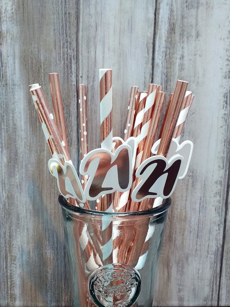 Op de afbeelding: Een glazen pot gevuld met roze gouden gestreepte en gestippelde papieren rietjes met witte en zwarte "21" uitsnijdingen die aan de rietjes zijn bevestigd.