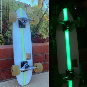 May include: A white skateboard with a green glow-in-the-dark stripe down the centre. The skateboard has black trucks and wheels.
