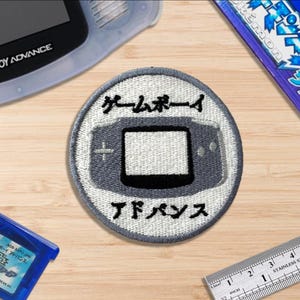 May include: An embroidered patch with a Game Boy Advance design in grey and black, with Japanese text. The patch is surrounded by a Game Boy Advance, a game cartridge, and a ruler, all on a wooden surface.