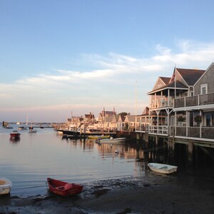 May include: A view of a waterfront town with houses built on stilts over the water. The houses are painted in shades of gray and white, with some red accents. There are several boats docked in the water, and the sky is a light blue with some clouds.