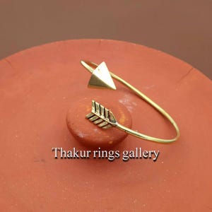 Può includere: Un bracciale a polsino color oro con un design a freccia. Il bracciale presenta una punta di freccia triangolare e un dettaglio di impennatura della freccia. Il testo "Thakur rings gallery" è visibile nella parte inferiore dell'immagine. Il bracciale è esposto su una superficie marrone rossastra.
