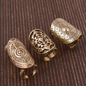 Puede incluir: Tres anillos ajustables de color dorado con intrincados diseños geométricos. Los anillos están hechos de metal y tienen un acabado pulido.
