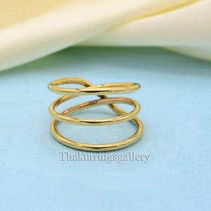 May include: A gold-colored ring with a unique design. The ring features three bands that are connected and intertwined. The ring is made of a shiny, metallic material and is set against a light blue background.