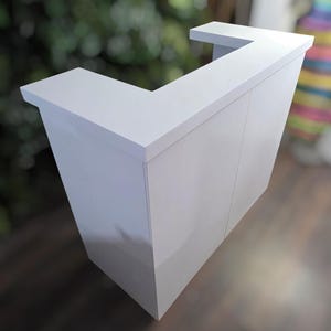 May include: A white, L-shaped bar with a glossy finish. The bar has a modern design with clean lines and a small overhang on the top surface. The bar is made of a solid material and appears to be suitable for indoor use.