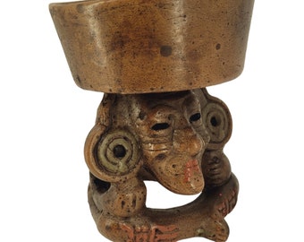 Puede incluir: Un recipiente de cerámica marrón con un cuenco cilíndrico ancho apoyado sobre una base esculpida con forma de rostro estilizado, orejas prominentes, adornos circulares en las orejas y detalles pintados en rojo.