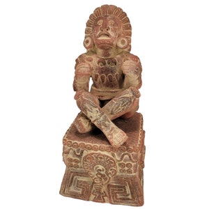 May include: A terracotta-colored sculpture of a seated figure with crossed legs. The figure has detailed facial features, ornate headdress, and decorative patterns on the chest and legs. The sculpture is seated on a square base with carved designs.