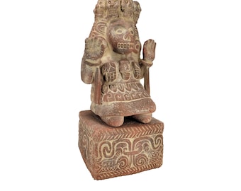May include: A terracotta sculpture of a seated figure with a skull-like face and a crown of skulls. The figure is wearing a patterned skirt and is seated on a square base with intricate carvings.