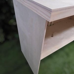 May include: A light-colored, unfinished wooden bench with a simple, modern design. The bench features a flat top and a lower shelf, with angled legs. The wood appears to be plywood. The bench is suitable for indoor or outdoor use.