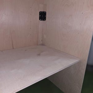 May include: Interior view of a wooden cabinet with a shelf. The cabinet is constructed from light-colored wood and features a black hinge. The shelf is positioned horizontally within the cabinet.