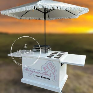 Może przedstawiać: Biały mobilny w&oacute;zek do serwowania z parasolem z frędzlami. W&oacute;zek zawiera r&oacute;żowy napis "Amayasweetbar Sweet & Delicious" oraz "Letty's Creations" w okręgu. Posiada wiele przegr&oacute;dek i obszar do serwowania.