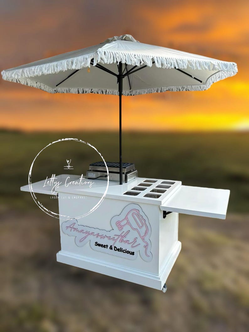 Może przedstawiać: Biały mobilny w&oacute;zek do serwowania z parasolem z frędzlami. W&oacute;zek zawiera r&oacute;żowy napis "Amayasweetbar Sweet & Delicious" oraz "Letty's Creations" w okręgu. Posiada wiele przegr&oacute;dek i obszar do serwowania.