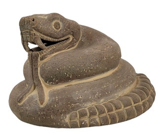 May include: A coiled, grey-brown snake sculpture with an open mouth, revealing teeth and a forked tongue. The snake's body is textured, with a detailed head and a patterned base. The sculpture is likely made of stone or a similar material.