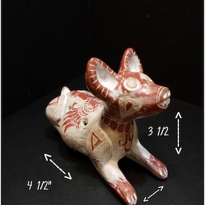 Può includere: Una statuetta di cane in ceramica dipinta di bianco e rosso. Il cane è sdraiato con la testa girata di lato. La statuetta è alta 9 cm, lunga 11,5 cm e larga 5 cm.