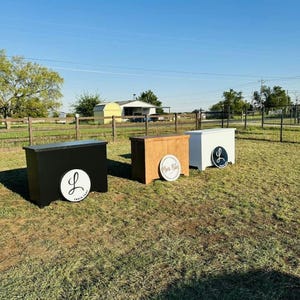 May include: Three wooden bar stands in a grassy field. One is black, one is brown, and one is white. Each has a circular logo on the front. The black bar has a white "L" logo, the brown bar has a "More Kicks" logo, and the white bar has a black "L" logo.