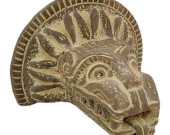 May include: A brown ceramic sculpture of a stylised animal head with a detailed design. The head has a large, open mouth with sharp teeth and a prominent nose. The sculpture is mounted on a wall.