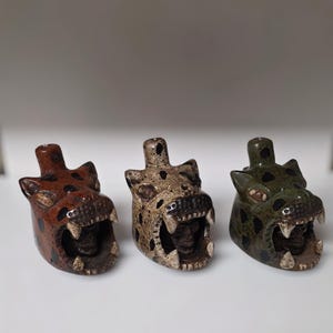 May include: Three ceramic jaguar head sculptures in shades of brown, tan, and green. Each jaguar has an open mouth revealing teeth and a skull-like interior. The sculptures have black spots and are set against a white background.