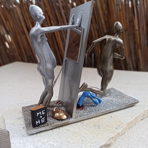 Op de afbeelding: Een zilveren en bronzen sculptuur van twee figuren, waarbij de ene de andere door een deur duwt. De deur is zilver geverfd en heeft een bruin paneel. Het beeld staat op een grijze basis met een zwart boek waarop "ME + ME" staat.