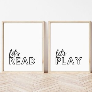 Puede incluir: Dos impresiones artísticas enmarcadas con las frases "let's READ" y "let's PLAY" en escritura negra y letras de bloque sobre un fondo blanco. Los marcos son de madera clara y el fondo es una pared blanca.