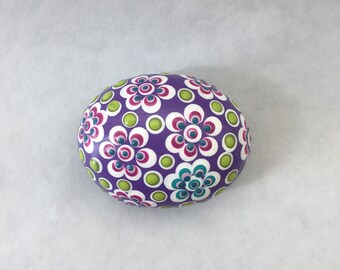 Puede incluir: Piedra pintada de morado con un patrón de flores blancas con centros rosas y azul verdoso y puntos verdes.