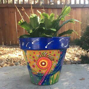 May include: A hand-painted flower pot with a vibrant design. The pot features a blue rim with white dots, and a colorful pattern of orange, pink, and green dots. The pot is filled with a green plant. The pot is sitting on a concrete surface.