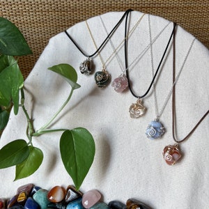 May include: Five necklaces with different colored stones wrapped in wire. The stones are pink, blue, brown, green, and gold. The necklaces are on a white fabric background.