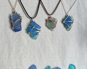 May include: Four necklaces with different colored sea glass pendants wrapped in wire. The pendants are blue, green, and light blue. The necklaces are on a white fabric background.