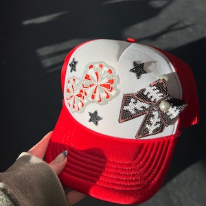 May include: Red and white trucker hat with a decorative design. The hat features a red brim and mesh back, with a white front panel adorned with pom-pom and star embellishments. A beaded bow with a pearl accent adds a touch of elegance.