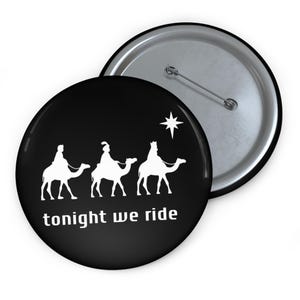 May include: A black button with a white graphic of three figures riding camels, a star, and the text "tonight we ride". The button is round and has a silver pin back.