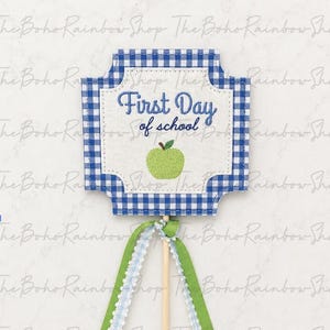 May include: A square-shaped sign with blue and white gingham borders, featuring the embroidered text First Day of school in blue and a green apple graphic. The sign is attached to a wooden stick adorned with light blue and green ribbons.