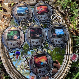 May include: A collection of metallic-colored keychains shaped like retro handheld game consoles. Each keychain features a different colorful screen image and a silver keyring. The keychains are arranged on a decorative mirror.