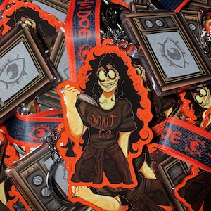 Puede incluir: Un llavero rojo y negro con un personaje de dibujos animados con el pelo largo y negro que sostiene un cuchillo. El personaje lleva una camiseta negra con el texto "Don't Go". El llavero también incluye una pequeña pantalla de televisión con una imagen estática.