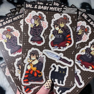 Könnte beinhalten: Eine Sammlung von Stickerbögen mit Illustrationen von Charakteren in verschiedenen Posen. Die Aufkleber haben einen braunen Hintergrund mit einem Rastermuster. Die Charaktere sind in schwarz-rot gestreiften Hemden dargestellt. Der Text "VAL & BABY MYERS" ist auf der Oberseite der Stickerbögen aufgedruckt.