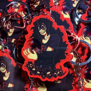 Puede incluir: Llaveros con una ilustración de dibujos animados de una figura con pelo largo y negro, una amplia sonrisa y sosteniendo una bola de cristal. El diseño tiene un borde rojo y está sobre un fondo de llaveros similares. Los llaveros tienen herrajes metálicos plateados y rojos.