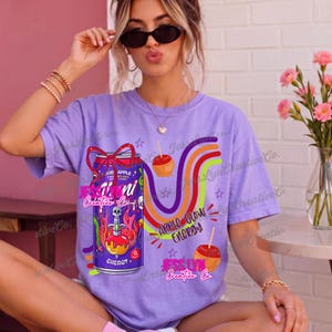Pode incluir: Uma t-shirt de manga curta roxo-claro com um gráfico de uma lata de bebida energética roxa com um laço vermelho, um crânio e uma adaga a atravessar uma maçã do amor, linhas onduladas coloridas e o texto Apple Glow Energy.