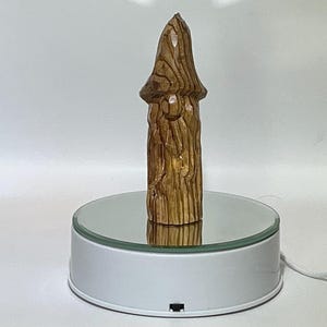 May include: A carved wooden sculpture of a mushroom with a textured surface and a light brown color. The mushroom is displayed on a rotating white platform with a mirrored top, showcasing its detailed craftsmanship.