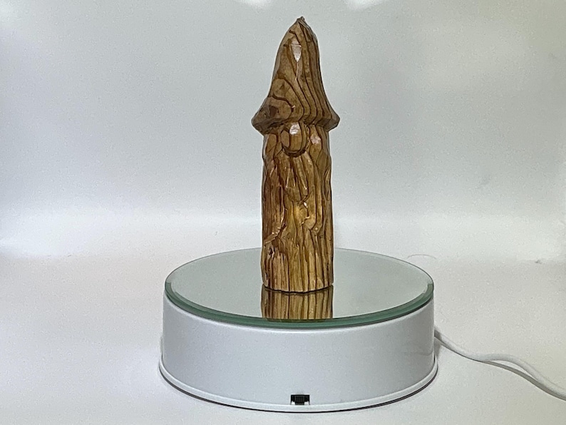 May include: A carved wooden sculpture of a mushroom with a textured surface and a light brown color. The mushroom is displayed on a rotating white platform with a mirrored top, showcasing its detailed craftsmanship.