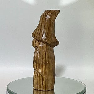 May include: A carved wooden sculpture of a gnome or similar figure, standing upright. The sculpture is a warm brown color, with visible wood grain and carved details. The figure has a pointed hat and a long, flowing beard.