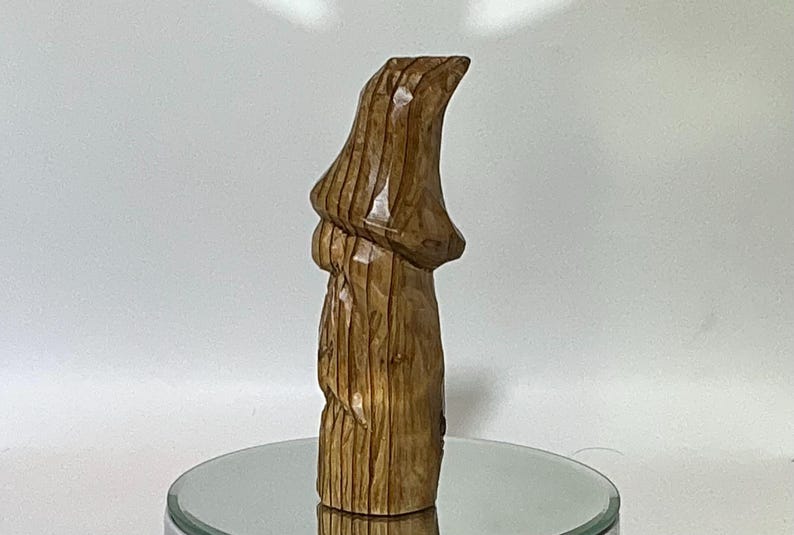 May include: A carved wooden sculpture of a gnome or similar figure, standing upright. The sculpture is a warm brown color, with visible wood grain and carved details. The figure has a pointed hat and a long, flowing beard.