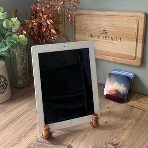 May include: A white tablet computer propped up on a wooden table with a copper coin stand. The tablet is displaying a black screen. The table is made of wood and has a light brown finish. There are other items on the table, including a wooden chopping board with the text "King of the Grill" and a stack of coasters.