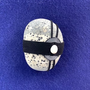 May include: A speckled gray stone adorned with a black band and a circular, layered gray and black design, centered with a white circular accent. The stone rests on a blue surface.