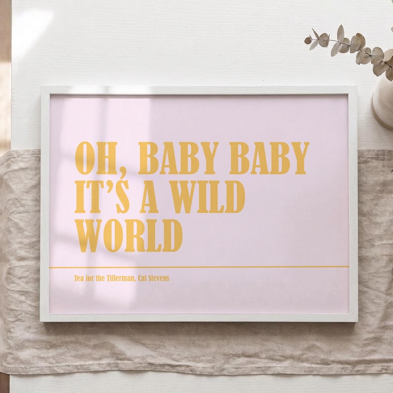 Op de afbeelding: Een ingelijste print met een lichtroze achtergrond en de tekst "OH, BABY BABY IT'S A WILD WORLD" in gouden letters. De tekst "Tea for the Tifferman, Cat Stevens" staat eronder. De lijst is wit.