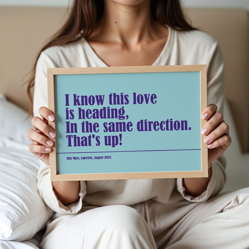 Op de afbeelding: Een ingelijste print met een lichtblauwe achtergrond en paarse tekst met de tekst: "I know this love is heading, In the same direction. That's up!" De print bevat ook de tekst "Olly Murs, Lakefest, August 2024."