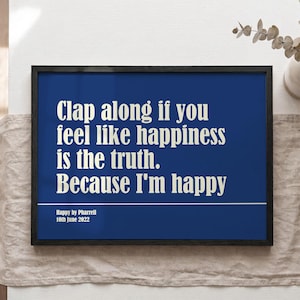 Op de afbeelding: Een ingelijste print met een marineblauwe achtergrond en witte tekst met de tekst "Clap along if you feel like happiness is the truth. Because I'm happy." Onder de tekst staat "Happy by Pharrell 10th June 2022."