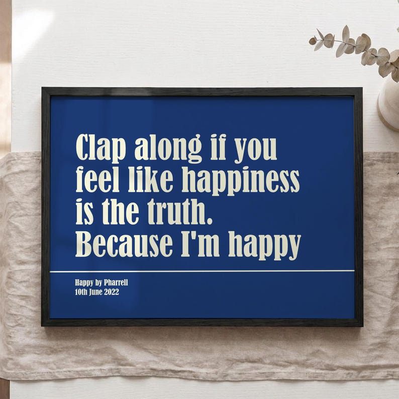 Op de afbeelding: Een ingelijste print met een marineblauwe achtergrond en witte tekst met de tekst "Clap along if you feel like happiness is the truth. Because I'm happy." Onder de tekst staat "Happy by Pharrell 10th June 2022."