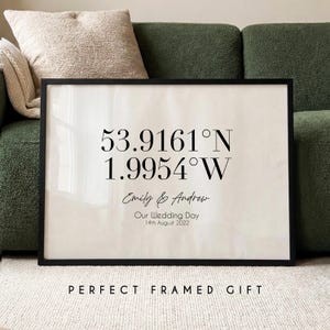 May include: A framed print with a black border displays coordinates, names, and a date. The text reads "53.9161°N 1.9954°W, Emily & Andrew, Our Wedding Day, 14th August 2022." The words "PERFECT FRAMED GIFT" are at the bottom.