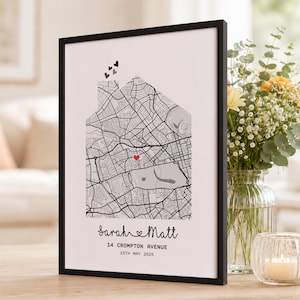 Op de afbeelding: Een ingelijste print met een zwart-witte stadsplattegrond in de vorm van een huis, met een rood hartje op een specifieke locatie. Boven de kaart staan drie kleine hartjes. Onderaan staat de tekst Sarah en Matt, 14 Crompton Avenue, 15 mei 2025.