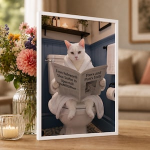 May include: A framed wall art print featuring a white cat wearing a white bathrobe, sitting on a toilet and reading a newspaper titled Paws and Purrs Daily with the headline From Failsworth Shoe Sniffer to Uppermill HoMEOwnner.