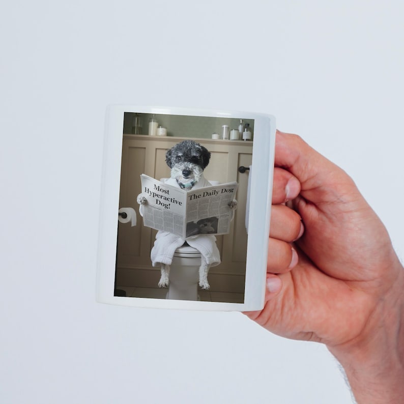 May include: A white ceramic mug featuring a printed image of a black and white dog wearing a white bathrobe whilst sitting on a toilet and reading a newspaper with the headline Most Hyperactive Dog and the title The Daily Dog printed on the front.