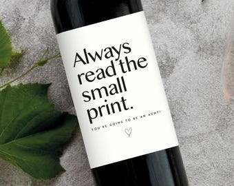 May include: A black wine bottle with a white label that reads "Always read the small print. You're going to be an aunt!" A green leaf is on the left side of the bottle. A glass of wine is in the background.